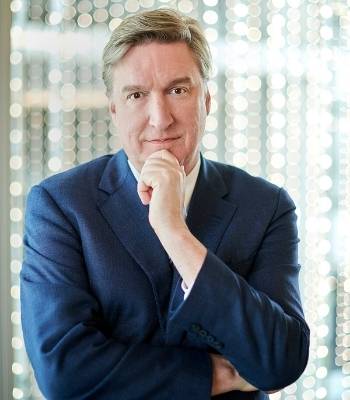 Byron Reese in blue suit with hand on chin and other arm folded standing in front of office window looking at camera